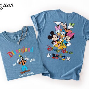 May include: Two blue denim-colored t-shirts. The left shirt features Goofy with the text Disneyland est. 1955 CALIFORNIA. The right shirt displays a group of Disney characters with the text Disney World 2026 and a silver chain necklace.