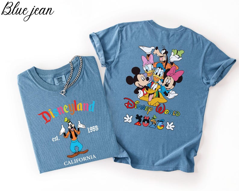 May include: Two blue denim-colored t-shirts. The left shirt features Goofy with the text Disneyland est. 1955 CALIFORNIA. The right shirt displays a group of Disney characters with the text Disney World 2026 and a silver chain necklace.