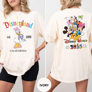 May include: An ivory t-shirt featuring a front graphic of Daisy Duck with the text Disneyland est. 1955 CALIFORNIA. The back graphic displays Mickey Mouse, Minnie Mouse, Donald Duck, Daisy Duck, Goofy, and Pluto with the text Disney World 2026.