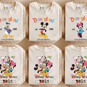 May include: Six off-white t-shirts featuring Disney character graphics. The top row shows Donald Duck, Mickey Mouse, and Minnie Mouse with the text Disneyland est. 1955 California. The bottom row displays a group of characters with Disney World 2026.