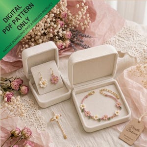 Puede incluir: Un conjunto de joyería de ganchillo que incluye un par de pendientes, una pulsera y un anillo con motivos florales, todos con pequeñas flores blancas y rosas. Los artículos se presentan en dos joyeros de terciopelo color crema con herrajes dorados.