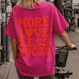 Könnte beinhalten: Ein leuchtend pinkes Kurzarm-T-Shirt mit dem orangefarbenen Text MORE LOVE LESS FUCKS, wobei ein Herz das O in LOVE ersetzt. Das T-Shirt wird mit einer Radlerhose mit Leopardenmuster und schwarzen Sandalen kombiniert, stehend neben einem Fahrrad.