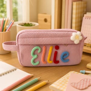 Op de afbeelding: Een lichtroze corduroy etui met de naam Ellie in kleurrijke, verhoogde, gebreide letters. Het etui is voorzien van een handvat aan de zijkant en een kleine, witte, pluche bloemversiering aan de rechterbovenhoek van het voorpaneel.