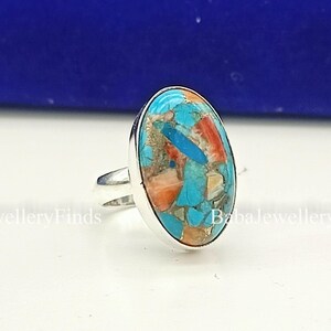 May include: A silver ring featuring an oval-shaped inlay composed of crushed turquoise, coral, and tan stone fragments. The metal band is polished and smooth, holding the multi-coloured mosaic stone setting securely in a simple, clean bezel frame.