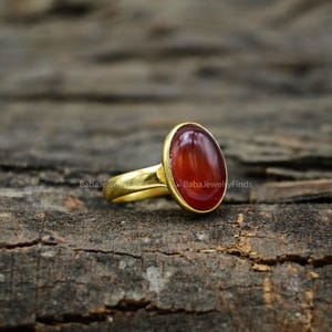 Può includere: Un anello color oro con una pietra preziosa cabochon di forma ovale, di un rosso aranciato intenso, incastonata in una semplice montatura in metallo. Il gambo dell'anello è liscio e lucido, con una finitura metallica calda.