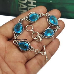 May include: A silver bracelet featuring teardrop-shaped, light blue gemstones. Each gem is set in a decorative silver frame, connected by silver links. The bracelet has a toggle clasp for secure fastening. The bracelet is displayed on a hand.