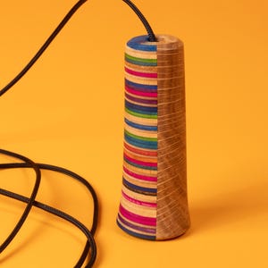 Könnte beinhalten: Ein zylindrischer Zugschaltergriff mit geteiltem Design. Eine Hälfte besteht aus gestapelten, mehrfarbigen horizontalen Schichten in Blau-, Grün-, Orange- und Rosatönen, während die andere Hälfte aus natürlicher Holzmaserung besteht. Ein schwarzes Kabel ist oben befestigt.