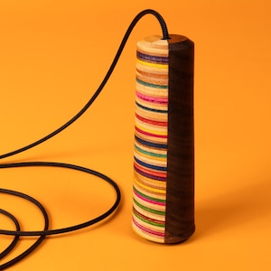 May include: A cylindrical pendant hanging from a black cord. One half of the cylinder is made of stacked, multi-coloured layers of wood in shades of pink, blue, yellow, and natural wood, while the other half is a solid, dark brown wood grain.