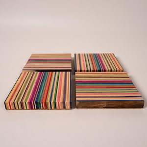 May include: Four square coasters made from stacked layers of multi-coloured wood. Each coaster features a distinct pattern of thin, vibrant horizontal and vertical stripes in shades of blue, red, yellow, green, and pink, with a natural wood base.