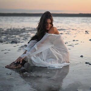 Woman sitting in shallow water wearing a sheer white off-shoulder tencel dress with puff sleeves during sunset.