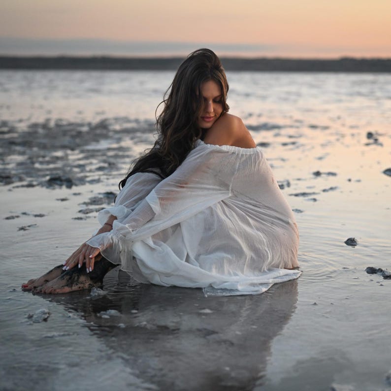 Woman sitting in shallow water wearing a sheer white off-shoulder tencel dress with puff sleeves during sunset.