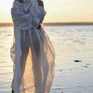 Close-up of a sheer white off-shoulder tencel coverup dress with puff sleeves on a background of water at sunset