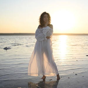 Woman in a sheer white off-shoulder boho dress with puff sleeves standing in shallow water during sunset.