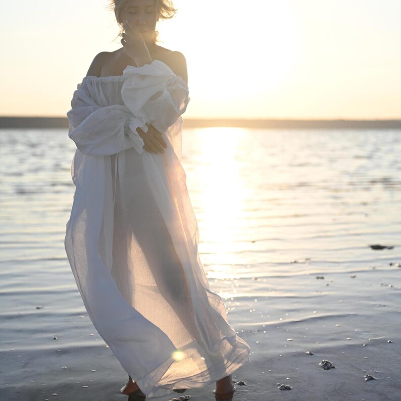 Front view of a woman posing in water wearing a flowing sheer white tencel off-shoulder boho dress at sunset