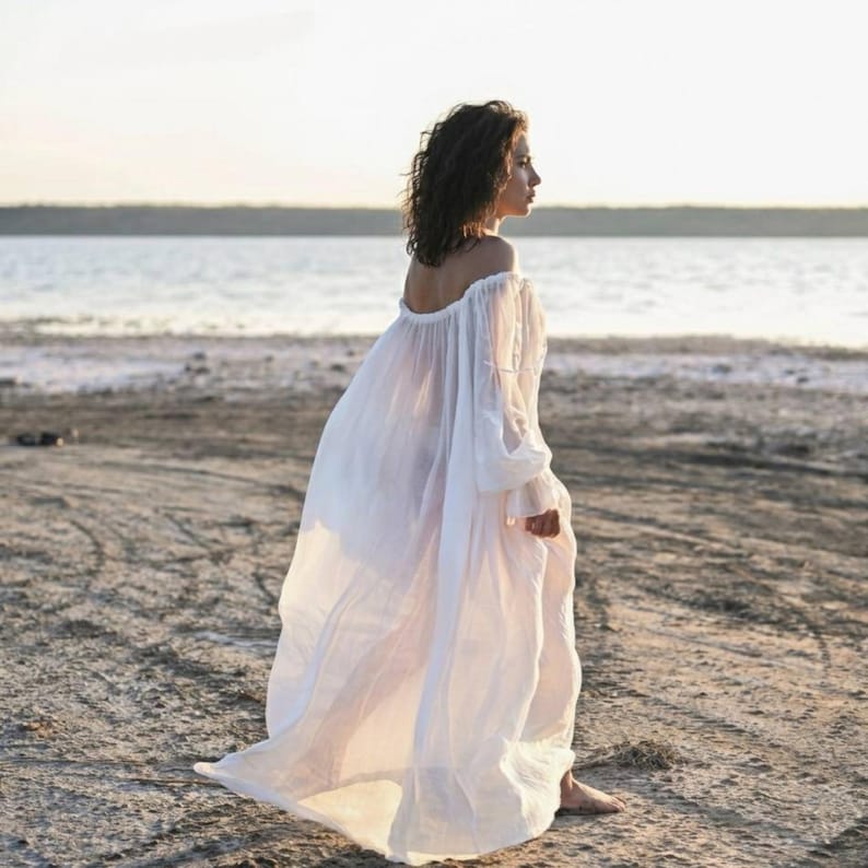 Side view of a woman walking on a sandy beach in a long sheer white tencel off-shoulder maxi dress.