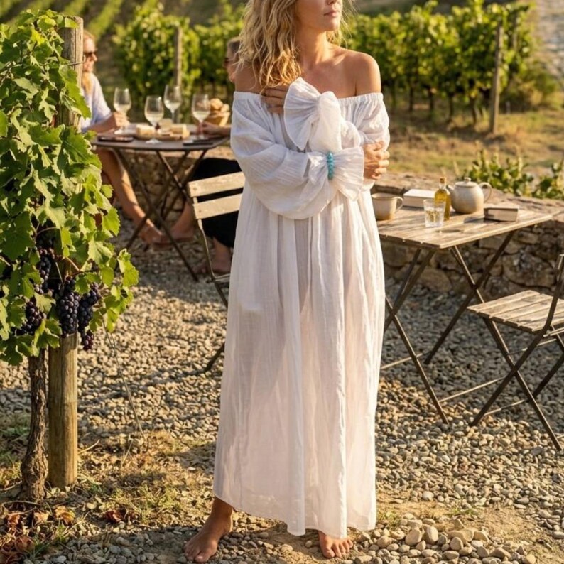 Woman standing barefoot in a vineyard wearing a white off-shoulder boho maxi dress with puff sleeves and turquoise bracelet.