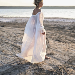 Side view of a woman walking barefoot on a shore in a flowing sheer white off-shoulder maxi tencel dress.