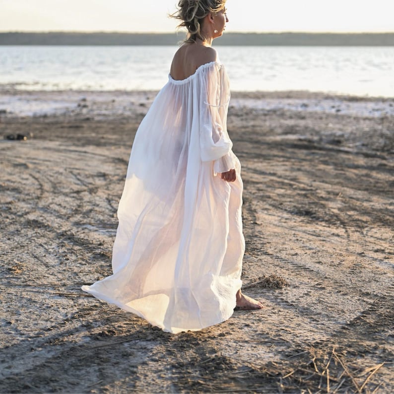 Side view of a woman walking barefoot on a shore in a flowing sheer white off-shoulder maxi tencel dress.