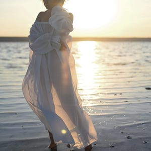 Full-length shot of a flowing sheer white tencel boho dress with puff sleeves standing in water at sunset.
