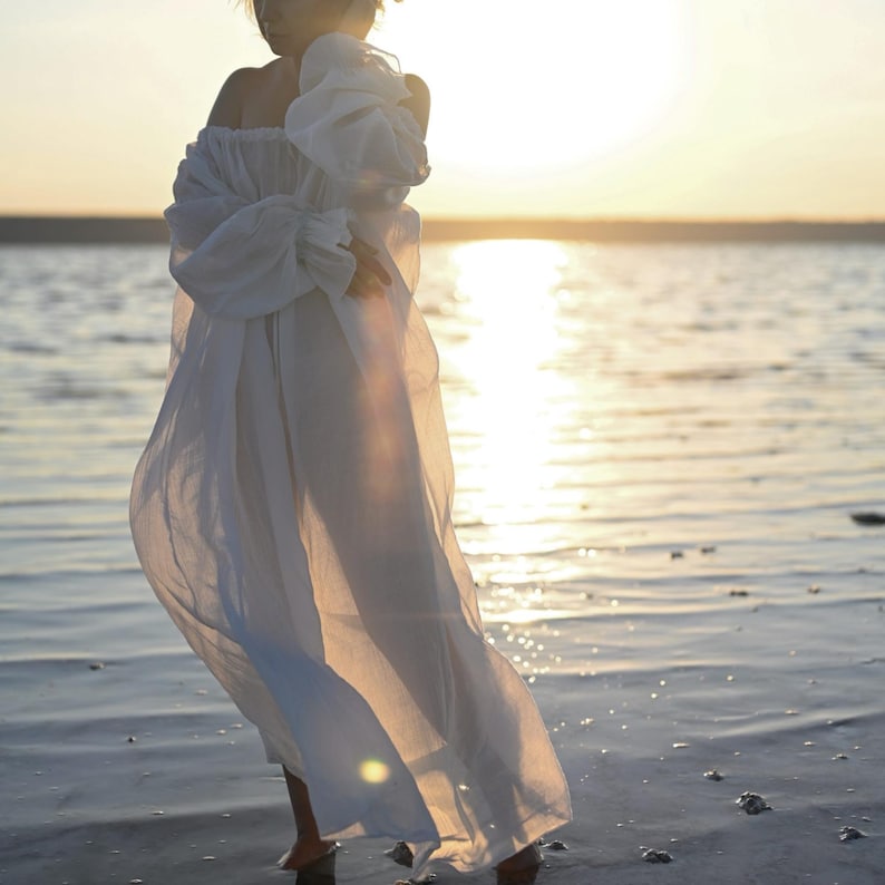 Full-length shot of a flowing sheer white tencel boho dress with puff sleeves standing in water at sunset.
