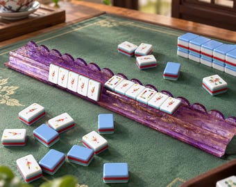 Puede incluir: Un juego de mahjong con fichas blancas que muestran ilustraciones de cócteles coloridos y reversos azules, sobre una superficie de fieltro verde. Un soporte largo de resina morada con remolinos dorados sostiene una fila de fichas, otras están dispersas.