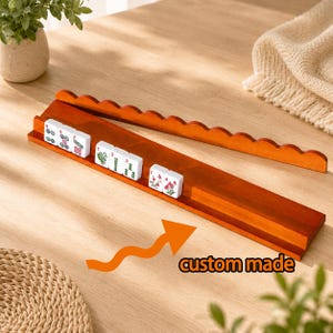 May include: A long, rectangular wooden mahjong tile rack with a scalloped top edge and a grooved ledge for holding tiles. Several white mahjong tiles with red, green, and black symbols are arranged on the rack. The text custom made is visible.