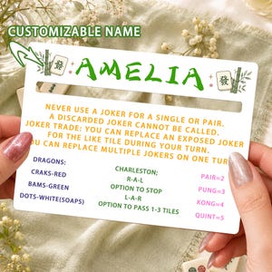 Può includere: Una scheda rettangolare bianca di riferimento per il Mahjong con il nome AMELIA in testo verde, affiancato da illustrazioni di bambù e tessere. La scheda include regole in testo giallo sui jolly e elenca categorie per draghi e Charleston.