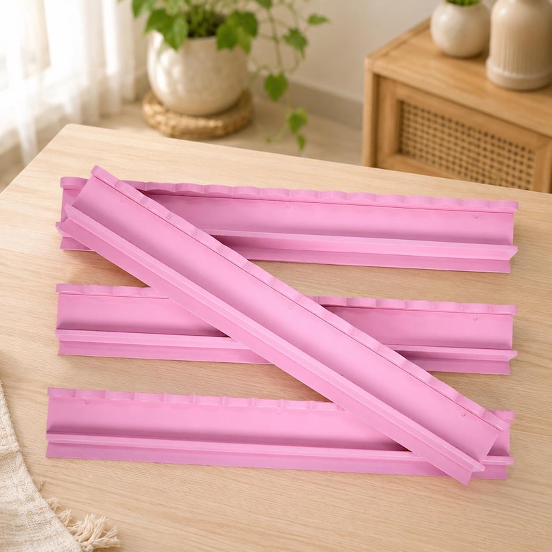 Puede incluir: Tres moldes de silicona largos de color rosa claro con un canal semicil&iacute;ndrico a lo largo del centro. Cada molde presenta un borde festoneado en la parte superior y una base plana, dise&ntilde;ados para crear formas redondeadas uniformes.