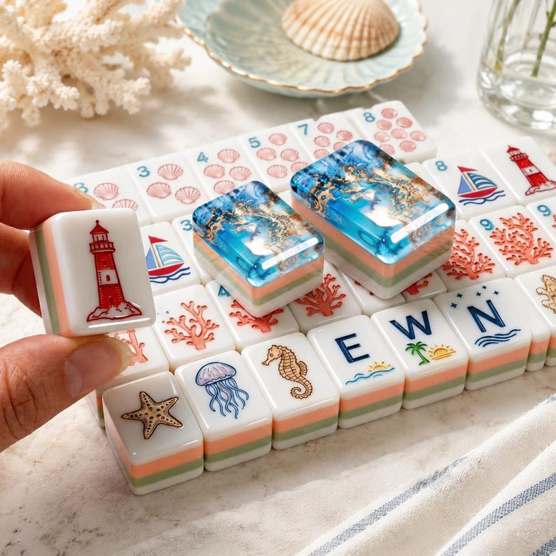 May include: A set of white mahjong tiles featuring nautical and ocean-themed illustrations including a red lighthouse, a sailboat, a starfish, a jellyfish, a seahorse, coral, and the letters E, W, and N, with two tiles displaying blue resin.