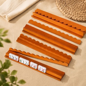 Könnte beinhalten: Ein Set aus vier langen, rechteckigen Holzleisten mit gewellter Oberkante, die zum Halten von Spielsteinen dienen. Eine Leiste enthält eine Reihe weißer, rechteckiger Spielsteine mit grünen und roten Blumenmotiven sowie numerischen Symbolen.