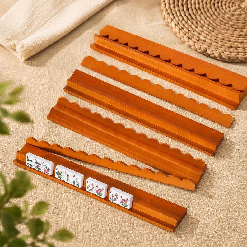 May include: A set of four long, rectangular wooden racks with a scalloped top edge, designed for holding game tiles. One rack contains a row of white rectangular tiles featuring green and red floral and numerical symbols in a horizontal arrangement.