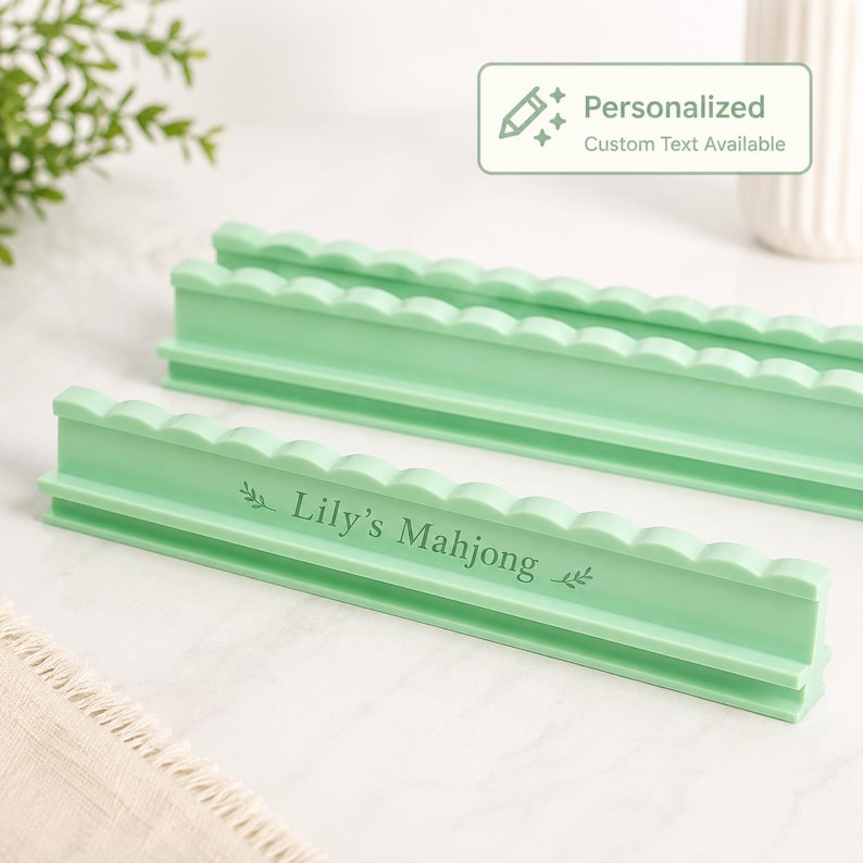 Puede incluir: Dos soportes para fichas de Mahjong de pl&aacute;stico verde claro con un borde superior festoneado. El soporte frontal presenta el texto grabado "Lily's Mahjong" flanqueado por peque&ntilde;os iconos de hojas. Un gr&aacute;fico rectangular muestra un icono de l&aacute;piz y el texto "Personalized Custom Text Available."