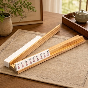 May include: Two light-colored wooden mahjong tile racks arranged at an angle. One rack holds white tiles with red Chinese characters, while the other rack is empty, showing a smooth, natural wood grain finish and a grooved design for holding tiles.