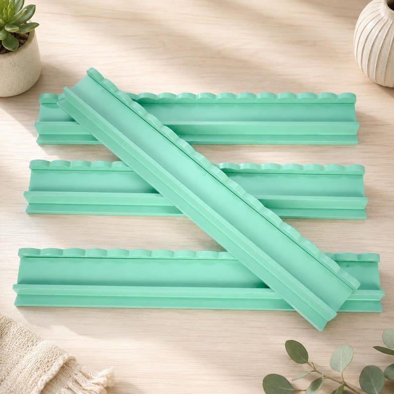 Puede incluir: Un conjunto de cuatro moldes de silicona de color verde menta, cada uno con un canal rectangular largo con un borde superior festoneado. Estos moldes flexibles de superficie lisa est&aacute;n dise&ntilde;ados para manualidades y dispuestos superpuestos.