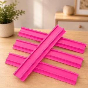 Puede incluir: Un conjunto de cuatro segmentos de pista de pl&aacute;stico rosa brillante, cada uno con un canal central y bordes elevados con un patr&oacute;n festoneado repetitivo en la parte superior, dispuestos en forma de cruz sobre una superficie de madera clara.