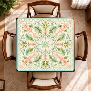 May include: A square tablecloth featuring a central compass rose motif surrounded by a symmetrical pattern of green palm fronds, pink hibiscus flowers, and seashells. The design is framed by a decorative border in shades of teal and cream.