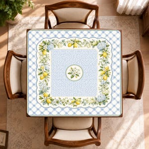 Puede incluir: Un mantel cuadrado con un panel central de patr&oacute;n azul claro y un motivo floral circular. Un borde de limones amarillos, flores blancas y racimos de hortensias azules enmarca el centro sobre un fondo de rejilla en blanco y azul.