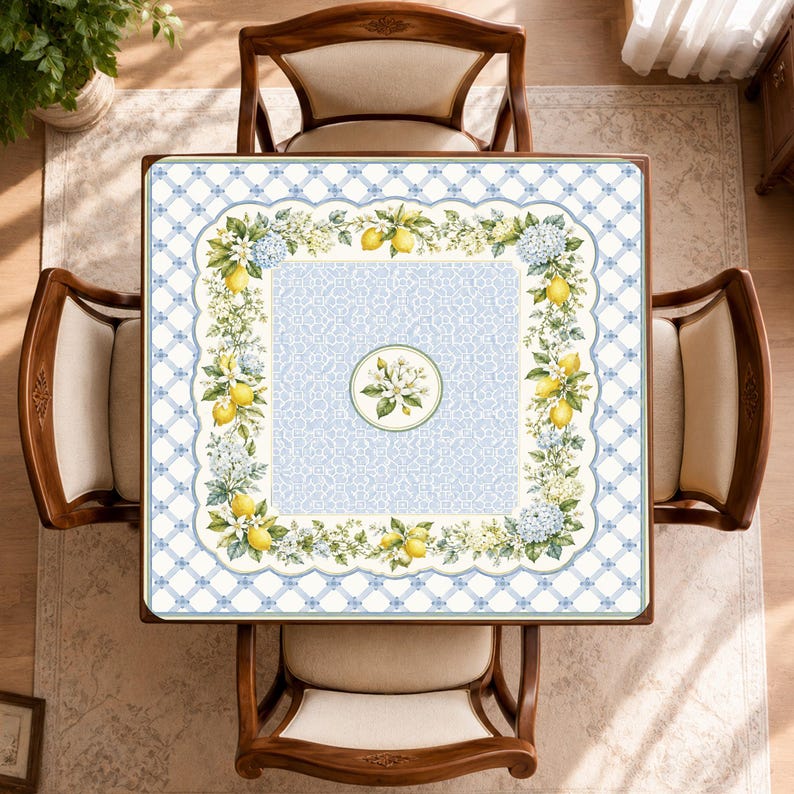 Puede incluir: Un mantel cuadrado con un panel central de patr&oacute;n azul claro y un motivo floral circular. Un borde de limones amarillos, flores blancas y racimos de hortensias azules enmarca el centro sobre un fondo de rejilla en blanco y azul.