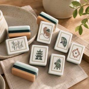 Pu&ograve; includere: Una collezione di tessere rettangolari da mahjong con facce color crema che presentano illustrazioni dettagliate di un leopardo, bamb&ugrave;, una pagoda, un ventaglio, un elefante e una cornice decorata, insieme a tessere con strisce verdi e beige.
