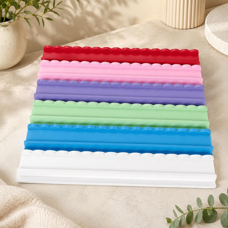 Puede incluir: Una pila de seis bandejas rectangulares de silicona, cada una con un borde superior festoneado. Las bandejas son de color rojo, rosa, morado, verde claro, azul y blanco, dispuestas en una columna vertical con una base plana y borde frontal.