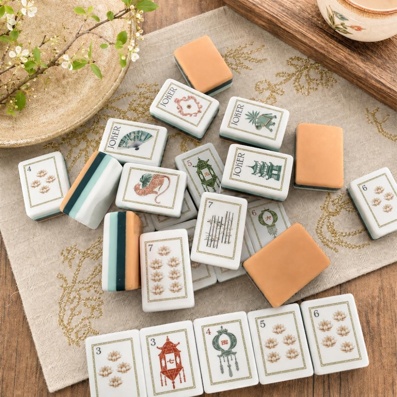 Pu&ograve; includere: Una collezione di tessere rettangolari da mahjong con varie illustrazioni tra cui pagode, ventagli, fiori e la parola Joker. Le tessere presentano facce bianche con disegni a inchiostro verde, rosso e nero e alcune hanno il retro beige.