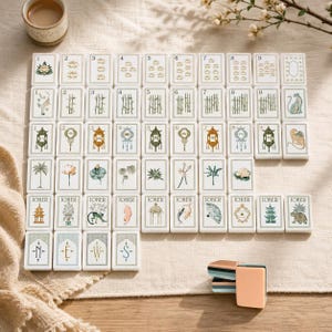 Pu&ograve; includere: Un set di tessere rettangolari da mahjong disposte in una griglia su una superficie in tessuto testurizzato. Le tessere bianche presentano varie illustrazioni tra cui fiori di loto, steli di bamb&ugrave;, lanterne e animali. Quattro tessere in basso mostrano le lettere N, E, W, S.
