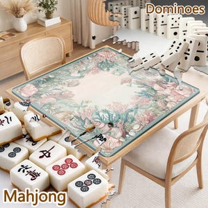 May include: A square game table featuring a light-colored surface with a printed border of seashells, coral, and seaweed. The table is surrounded by white mahjong tiles with black and red symbols and a row of white dominoes with black pips.