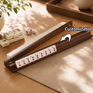 May include: Two dark brown wooden mahjong tile racks with a grooved design. One rack holds nine white mahjong tiles with red and black Chinese characters and numbers. The text "Customizable" is printed above the rack with a white arrow pointing down.