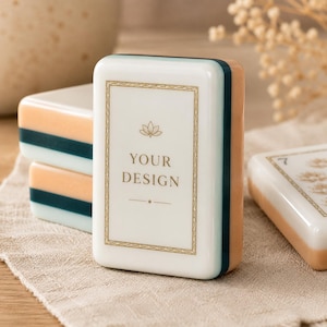Pu&ograve; includere: Una tessera rettangolare da mahjong con una faccia bianca che presenta un'icona di loto color oro, il testo "YOUR DESIGN" in oro e un bordo dorato. La tessera ha lati a strati nei colori bianco, verde ottanio scuro e pesca, finitura liscia.