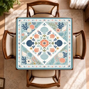 May include: A square tablecloth featuring a central diamond pattern filled with coral, navy blue, and light blue seashells, flowers, and leafy sprigs. The border displays a repeating motif of small orange and blue petals on a light blue background.