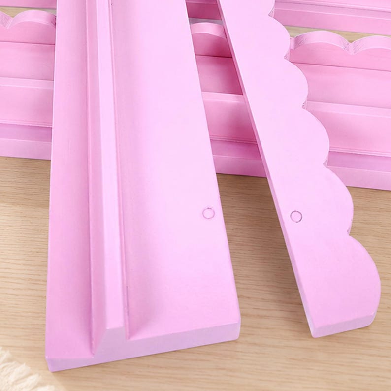 Puede incluir: Un conjunto de estantes de pared de madera de color rosa claro con un dise&ntilde;o de borde festoneado. Cada estante tiene una base plana con un borde frontal elevado y un panel trasero vertical. Las piezas est&aacute;n pintadas en rosa mate con orificios.