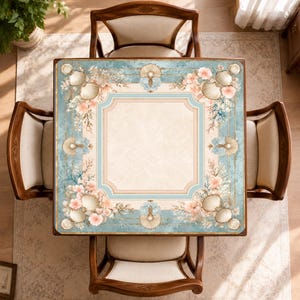 May include: A square table with a light beige center and a decorative border featuring blue tones, white seashells, and soft pink floral accents. The table is surrounded by four wooden chairs with light-colored upholstered seats and backrests.