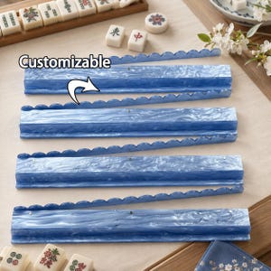 Puede incluir: Cuatro soportes de acrílico azul marmolado para fichas de mahjong con un borde superior festoneado. Cada soporte presenta un acabado liso y brillante con patrones de remolinos en azul claro y oscuro. El texto "Customizable" aparece con una flecha.