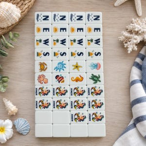 May include: A set of white rectangular mahjong tiles arranged in a grid. The tiles feature printed designs including the word NEWS with weather icons, various sea creatures like crabs and turtles, and illustrations of mermaids labeled as JOKER.